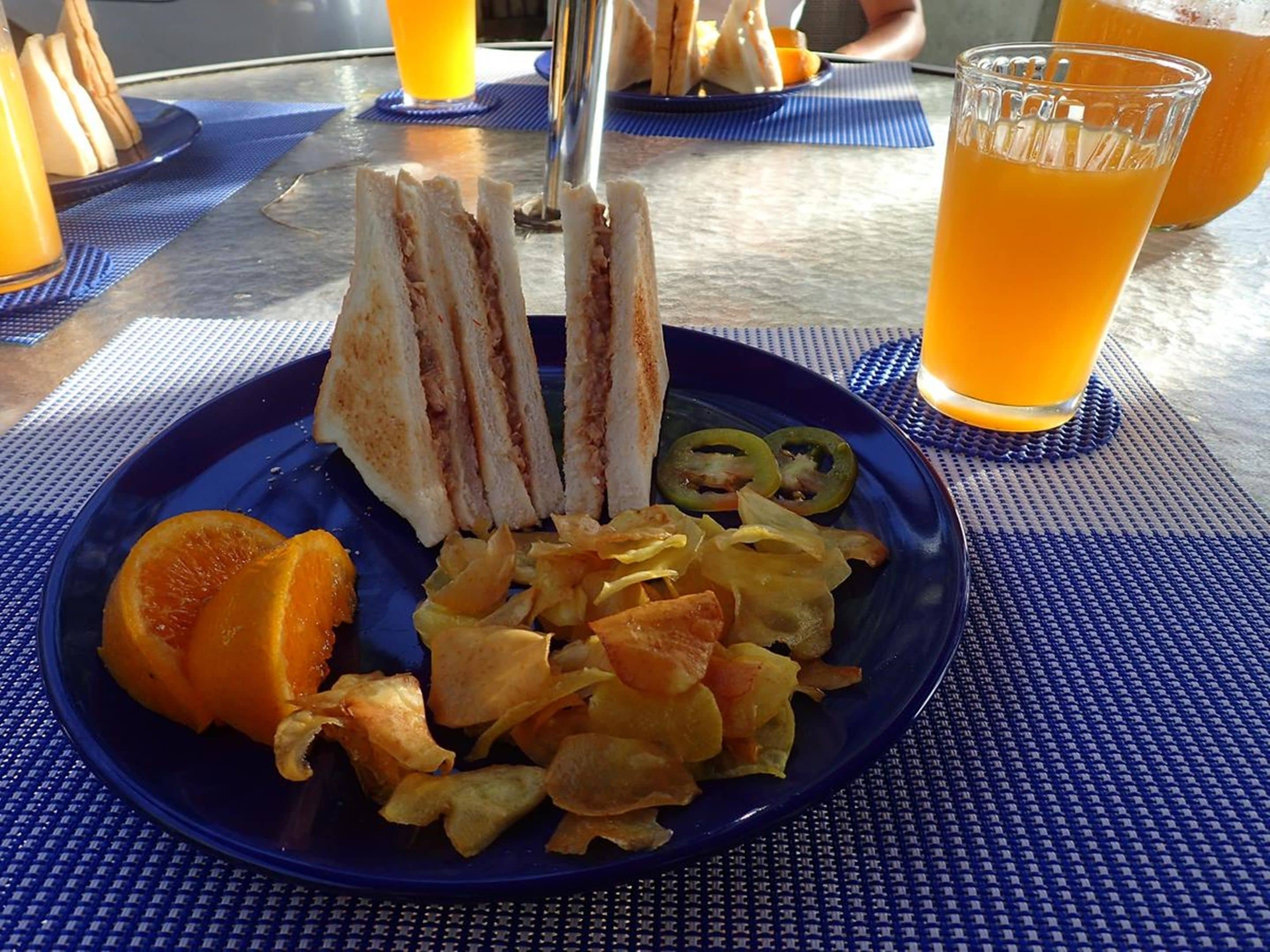 Un plato con sándwiches, jalapeños en rodajas y papas fritas, acompañado de un vaso de jugo de naranja.