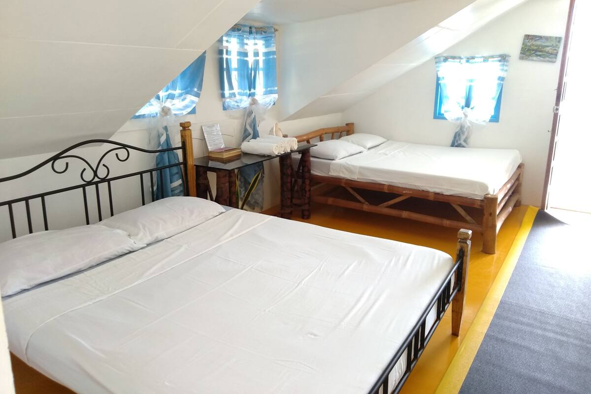 Ein Zimmer mit zwei Betten, einem kleinen Tisch und einem Fenster mit Vorhängen.