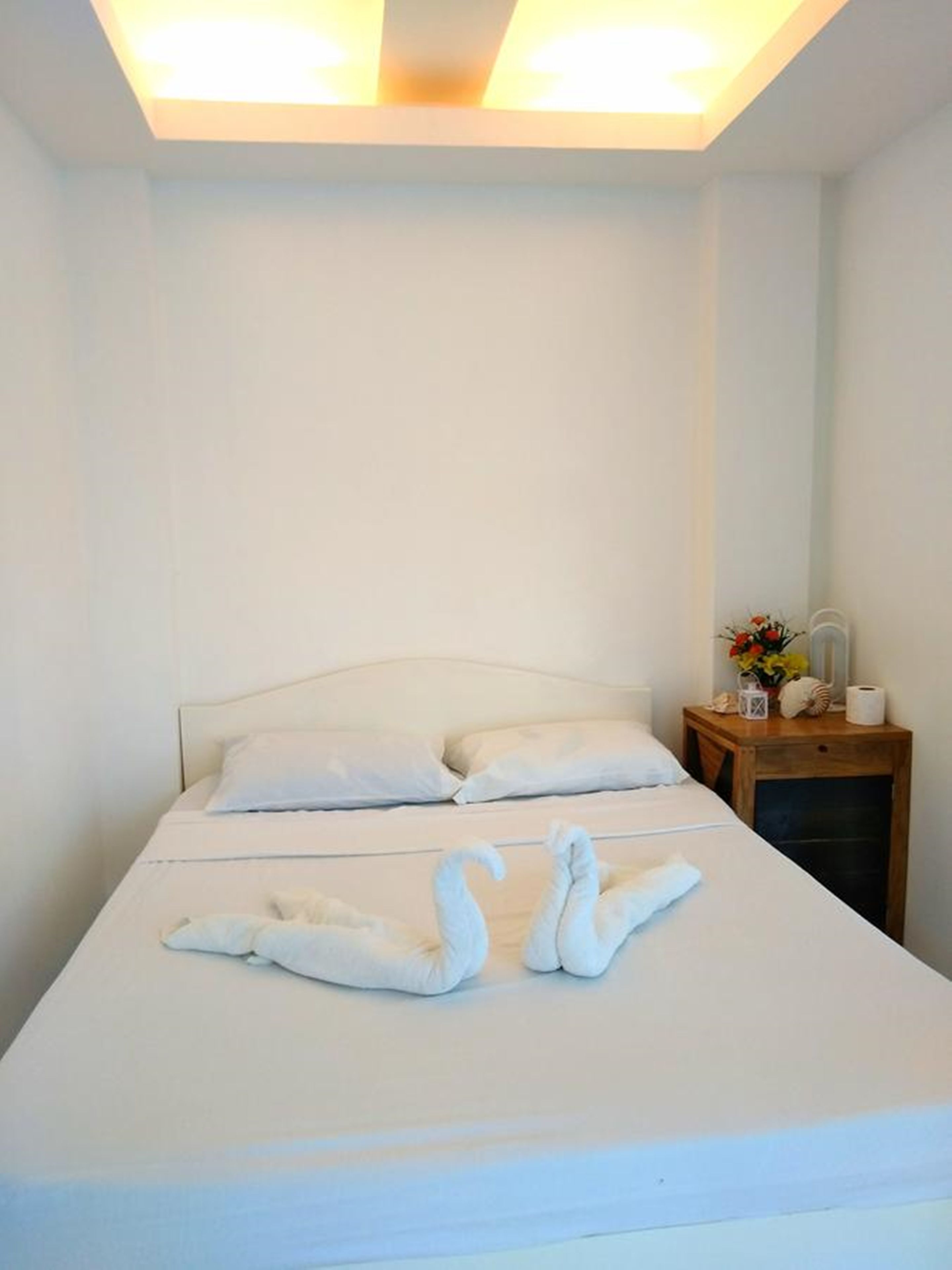 Una cama bien hecha con dos almohadas y dos toallas con forma de cisne. Junto a la cama, una mesita de noche de madera con un florero y una lámpara.