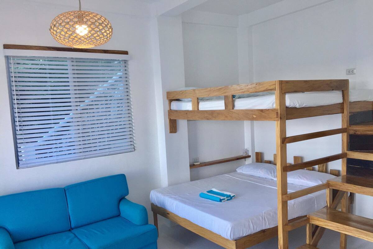 Ein Zimmer mit einem Etagenbett, einem blauen Sofa, einer Hängelampe und einem Fenster mit Jalousien.