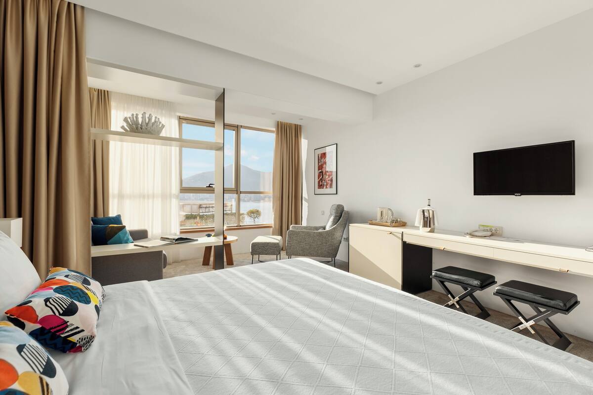 Ein Hotelzimmer mit Bett, Schreibtisch, Sessel und Fernseher, mit Blick auf die Stadt.