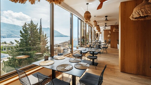 A restaurant with large windows offering a view of a lake and mountains. Tables are set with woven placemats and chairs. - Hotel Harmonia by Dukley (Budva)