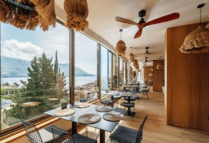 Un restaurante con amplios ventanales que ofrecen vistas a un lago y montañas. Las mesas están dispuestas con individuales tejidos y sillas.