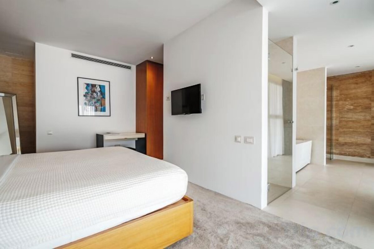 A modern bedroom with a bed, a flat-screen TV, and a bathroom visible through an open door.