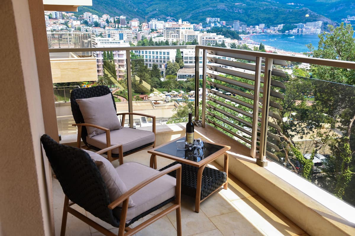 Ein Balkon mit zwei Korbstühlen, einem kleinen Tisch mit einer Flasche und Blick auf eine Küstenstadt.