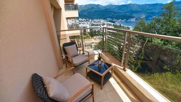A balcony with a view of a coastal city, a table with a bottle, and two chairs.