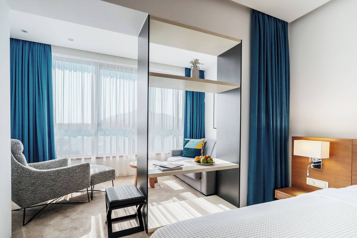 Ein modernes Hotelzimmer mit einem großen Bett, einer Sitzecke mit Stuhl und kleinem Tisch, einem Nachttisch mit Lampe sowie Blick auf die Stadt durch große Fenster.