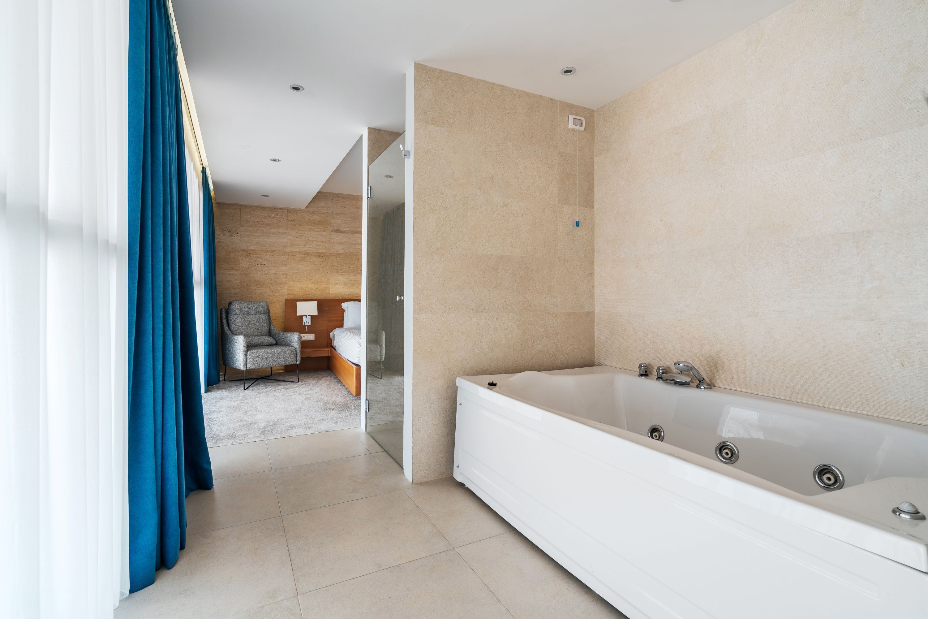 A modern bathroom with a large bathtub, a wooden floor, and a view into a bedroom with a bed and a chair.