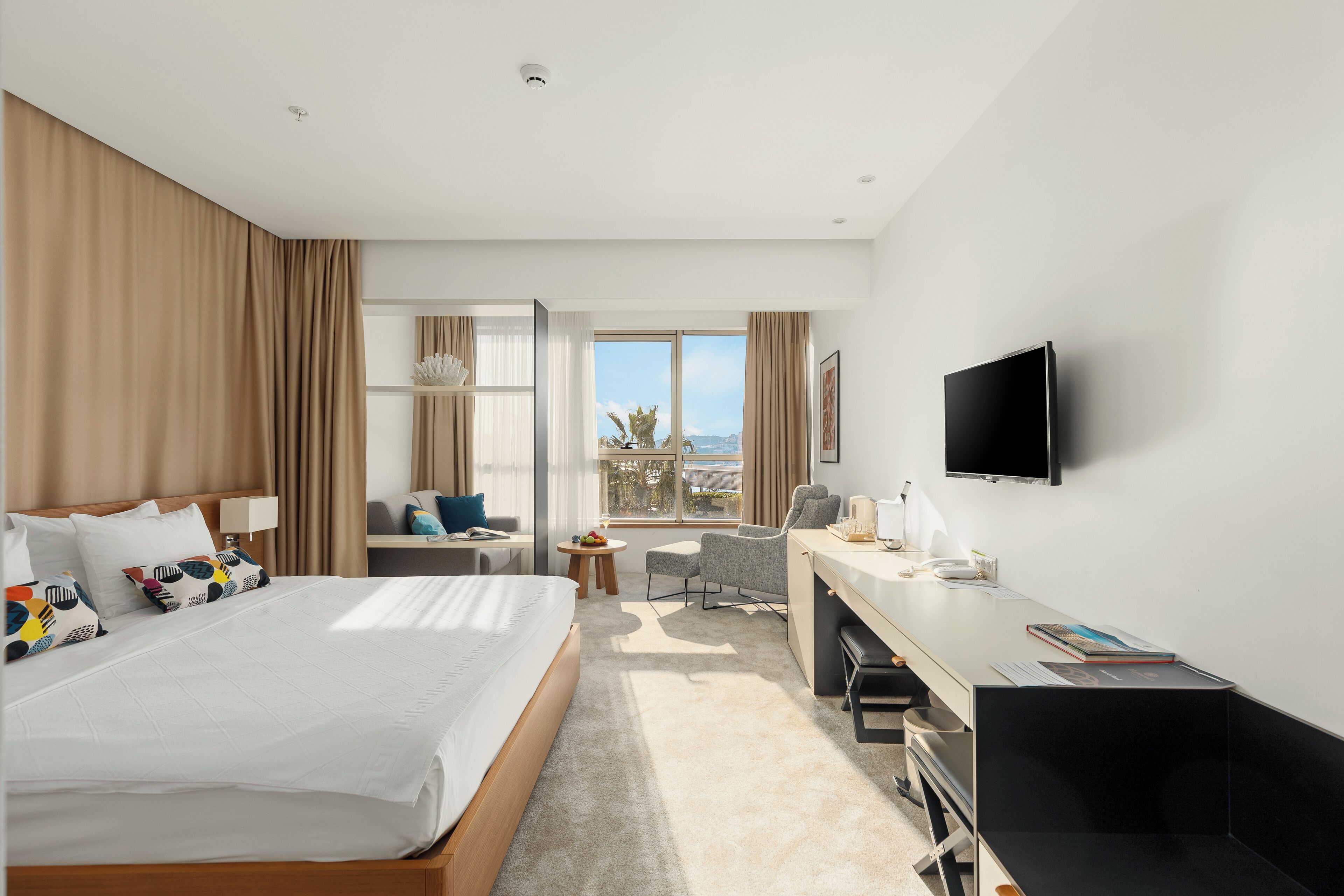 A hotel room with a large bed, a desk, a chair, a television, and a window with a city view.