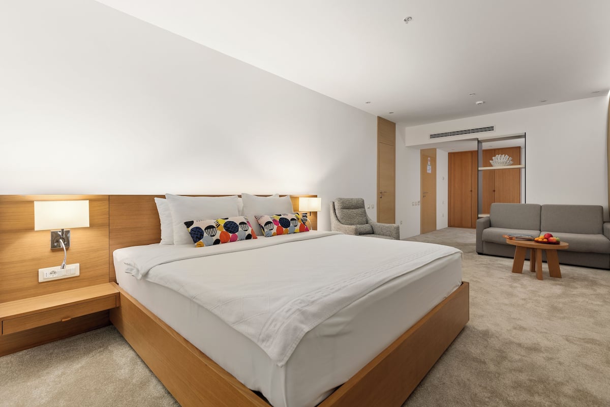 Ein modernes Hotelzimmer mit einem großen Bett, einem Nachttisch mit Lampe, einem grauen Sessel, einer grauen Couch und einem kleinen Holztisch mit Obst.