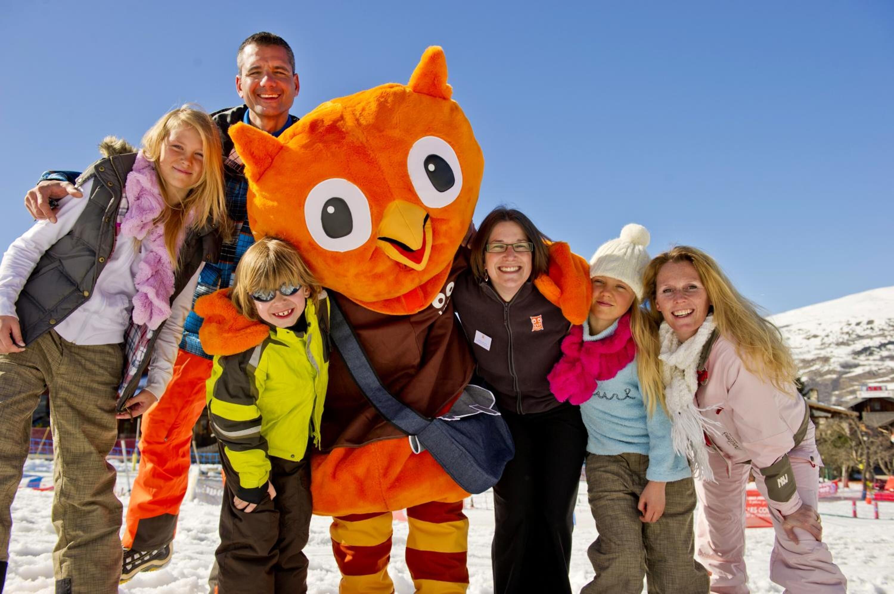 Eine Gruppe von Menschen posiert mit einer großen, orangefarbenen Maskotte in einer verschneiten Winterlandschaft.