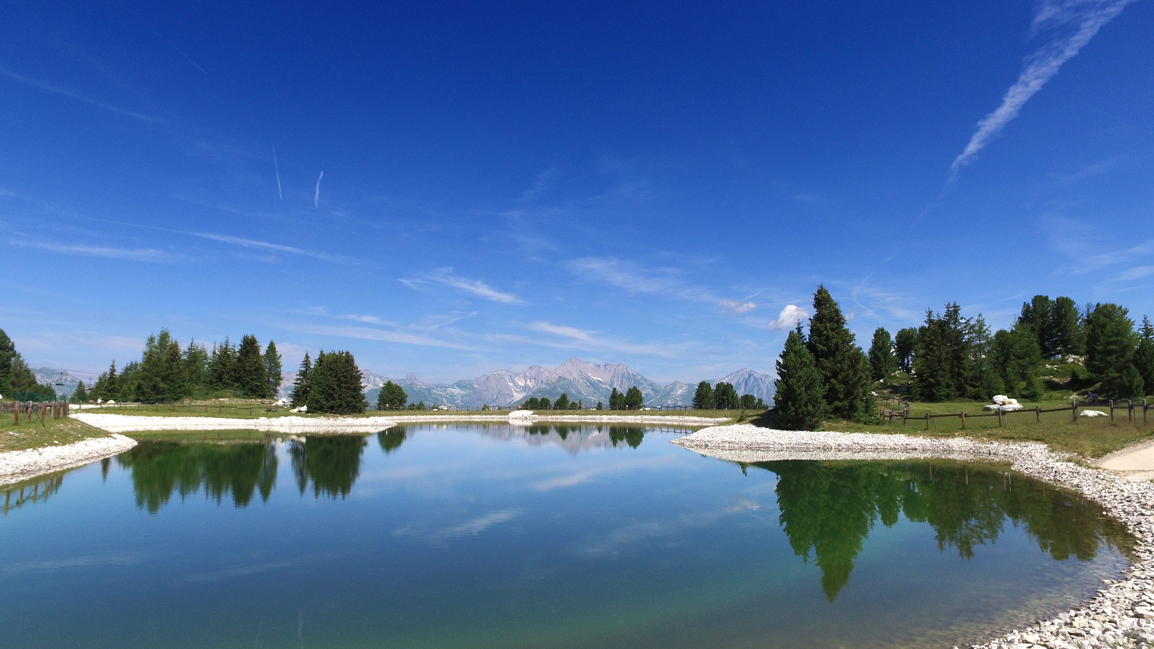 A tranquil lake with clear reflections, surrounded by a forest and mountains in the distance.