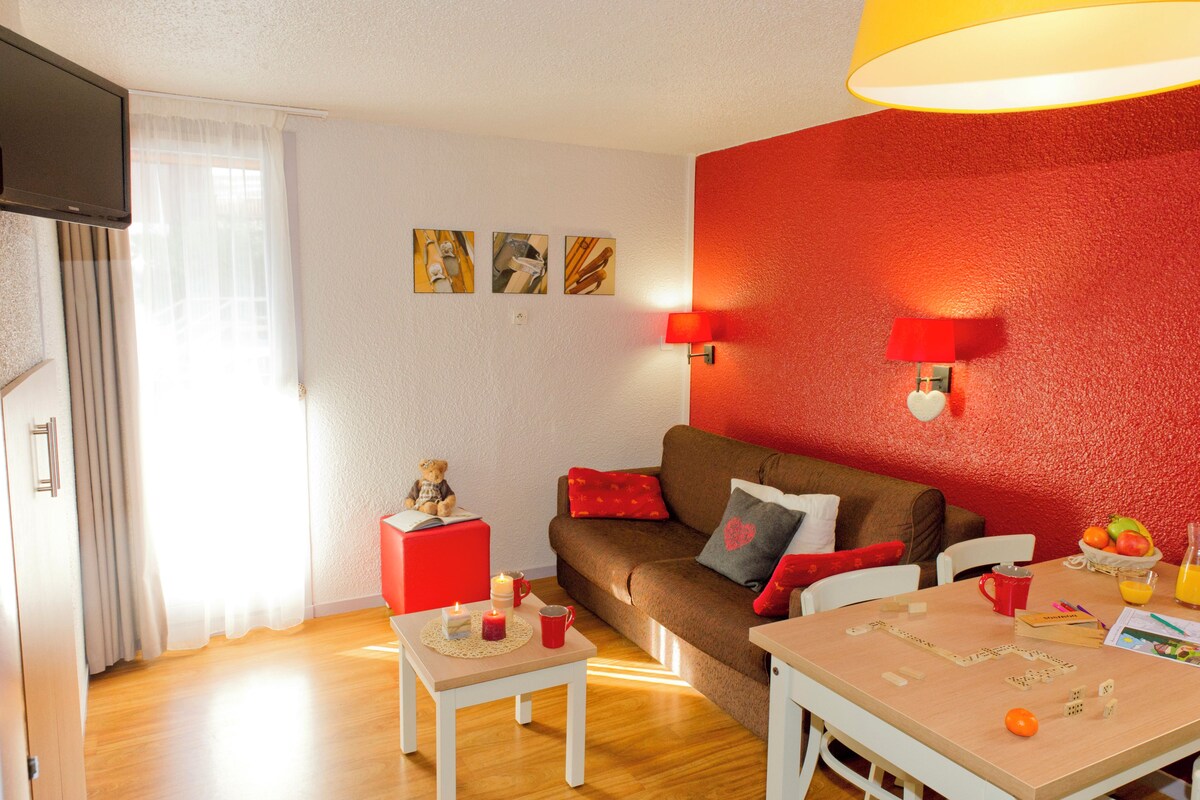 A cozy living room with a sofa, a small table with candles, and a dining area with a fruit bowl and a red lamp.