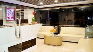 A modern hotel lobby with a reception desk, a sofa, a coffee table, and a decorative plant. - Hotel Bauji Palace (Udaipur)
