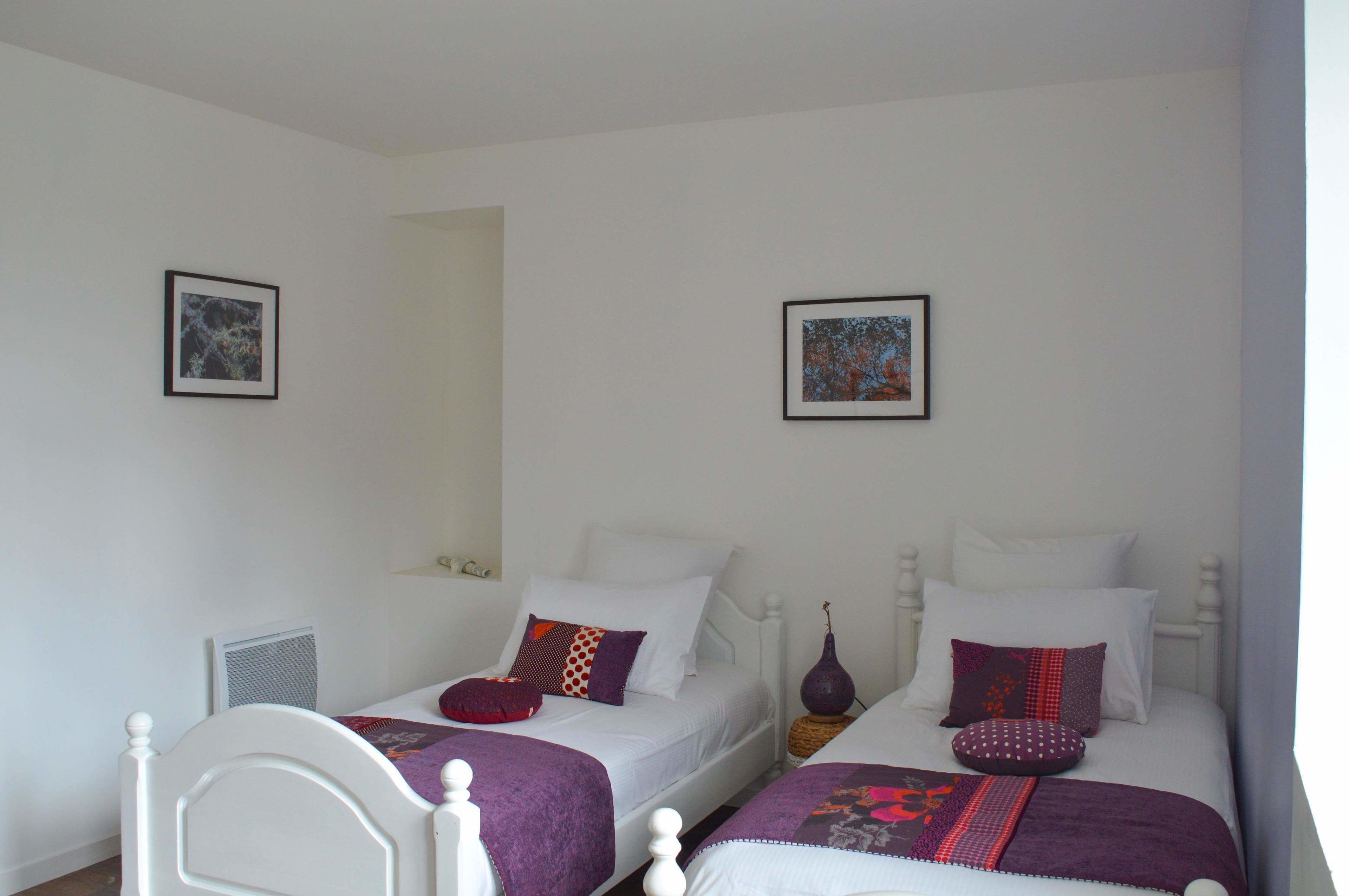 Une chambre avec deux lits simples, chacun doté d’une tête de lit blanche, d’une table de chevet et d’une lampe. Deux tableaux encadrés ornent le mur.
