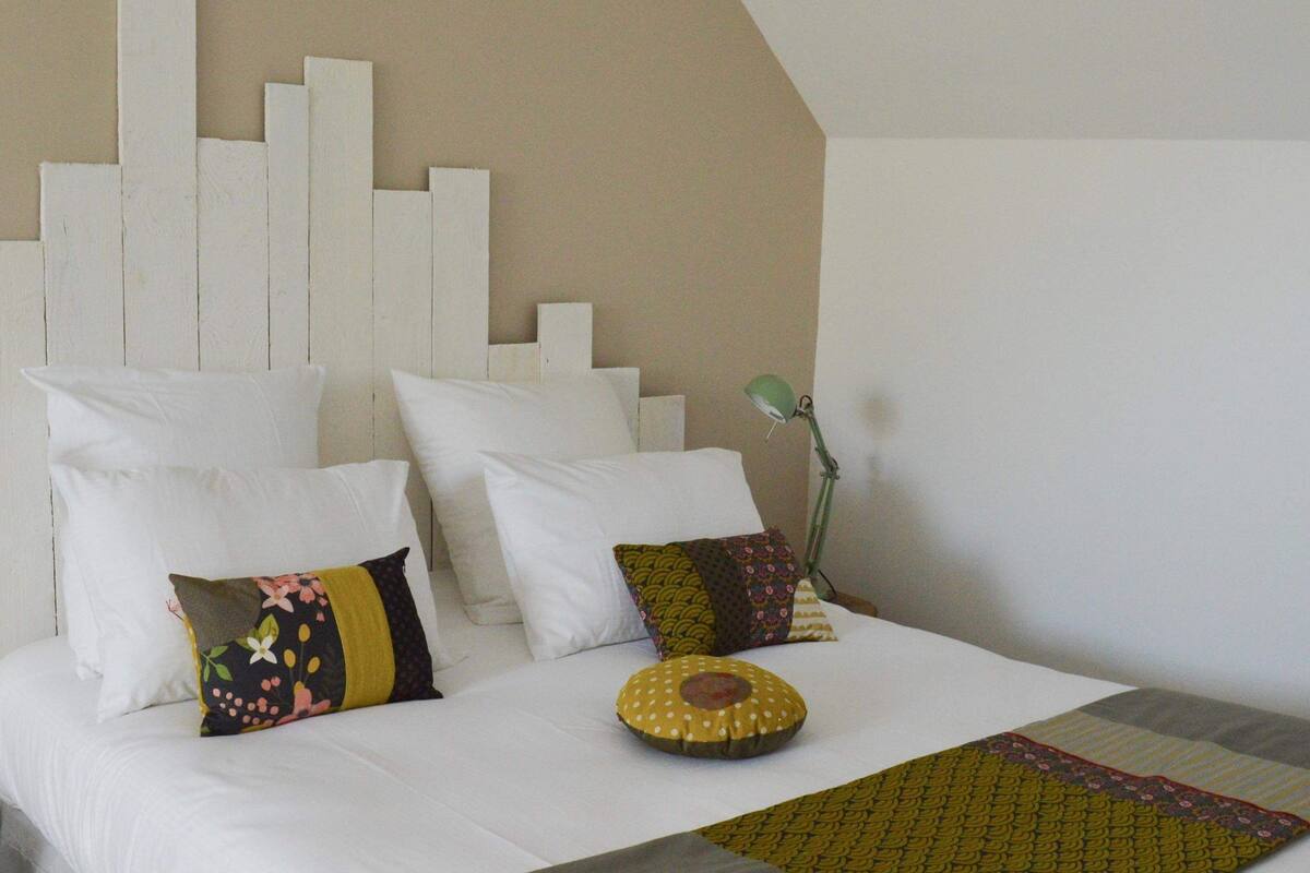 A neatly made bed with white bedding and multiple pillows, a green lamp on the bedside table, and a patterned throw blanket.