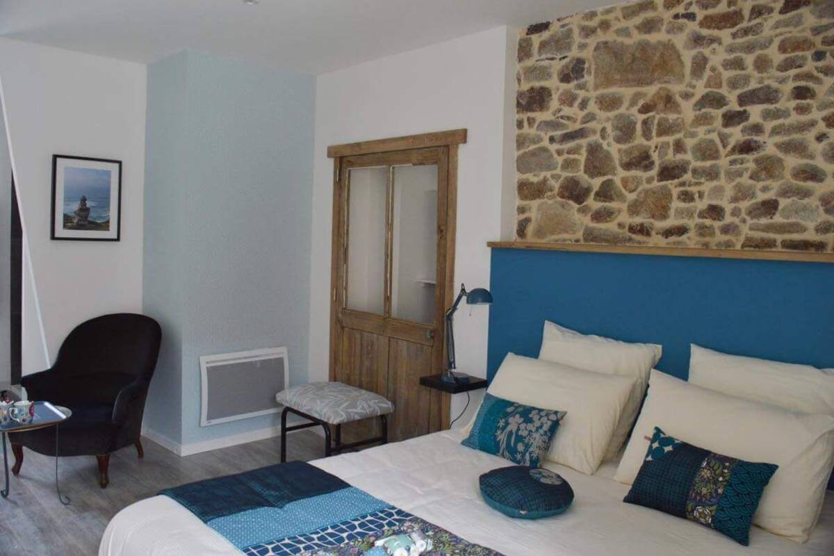 A bedroom with a stone wall, a bed with multiple pillows, a chair, a small table, and a picture on the wall.
