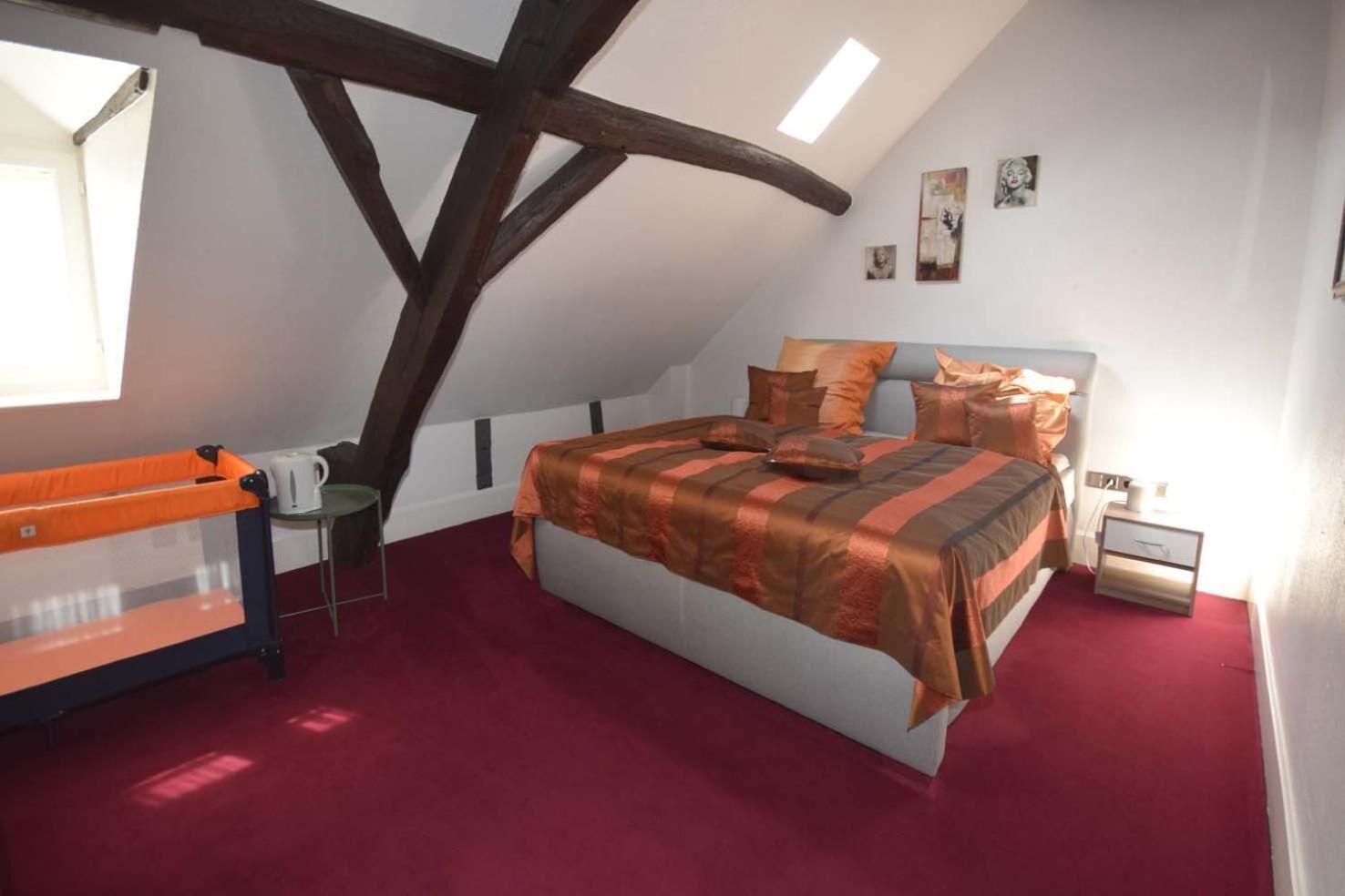 Ein Schlafzimmer mit schrägverdeckter Decke, einem Bett mit gestreifter Bettdecke, einem Nachttisch, einem kleinen Tisch mit Lampe und einem Fenster.