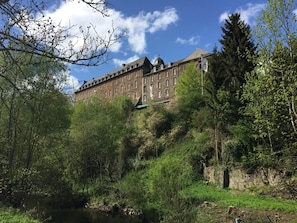 Un edificio grande e histórico con múltiples ventanas y una cúpula destacada, rodeado de vegetación exuberante y una pequeña extensión de agua.