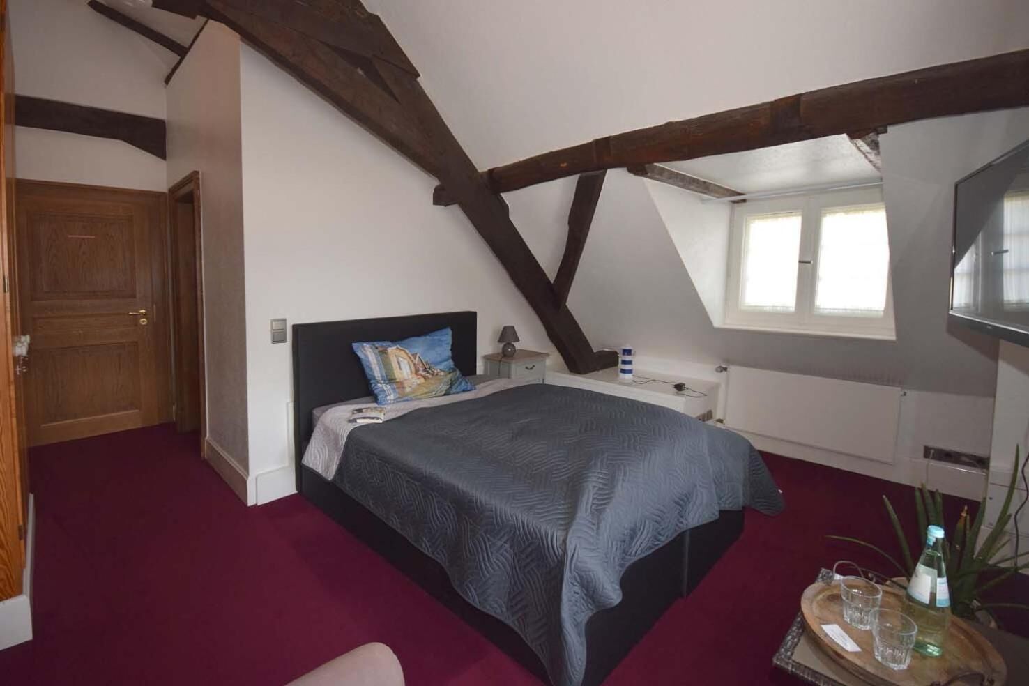 Een slaapkamer met een bed, een houten deur, een raam en een tafeltje met glazen en een fles.