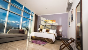 Une chambre d’hôtel moderne avec une grande fenêtre, un lit avec du linge de maison blanc, un canapé gris et une petite table.
