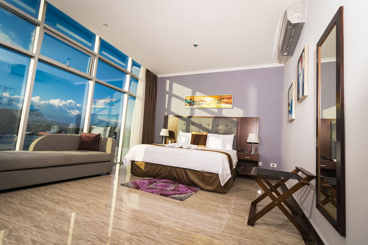 A modern hotel room with a large window, a bed with white linens, a grey sofa, and a small table.