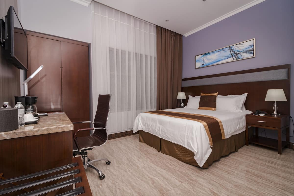 A hotel room with a bed, desk, chair, and a painting on the wall.