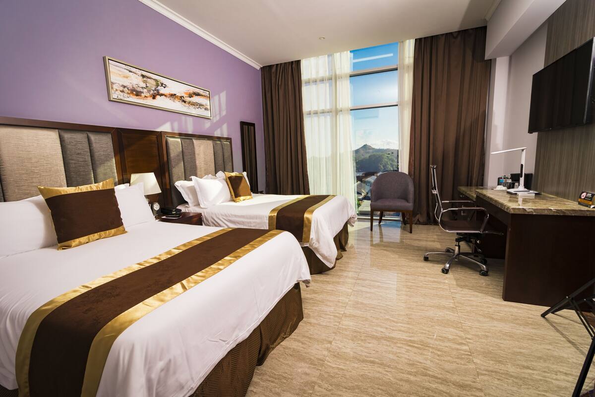 A hotel room with two beds, a desk, a chair, and a large window offering a mountain view.