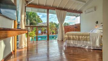 A spacious room with a wooden floor, a bed with white bedding, a TV, and a large sliding glass door leading to a pool area.