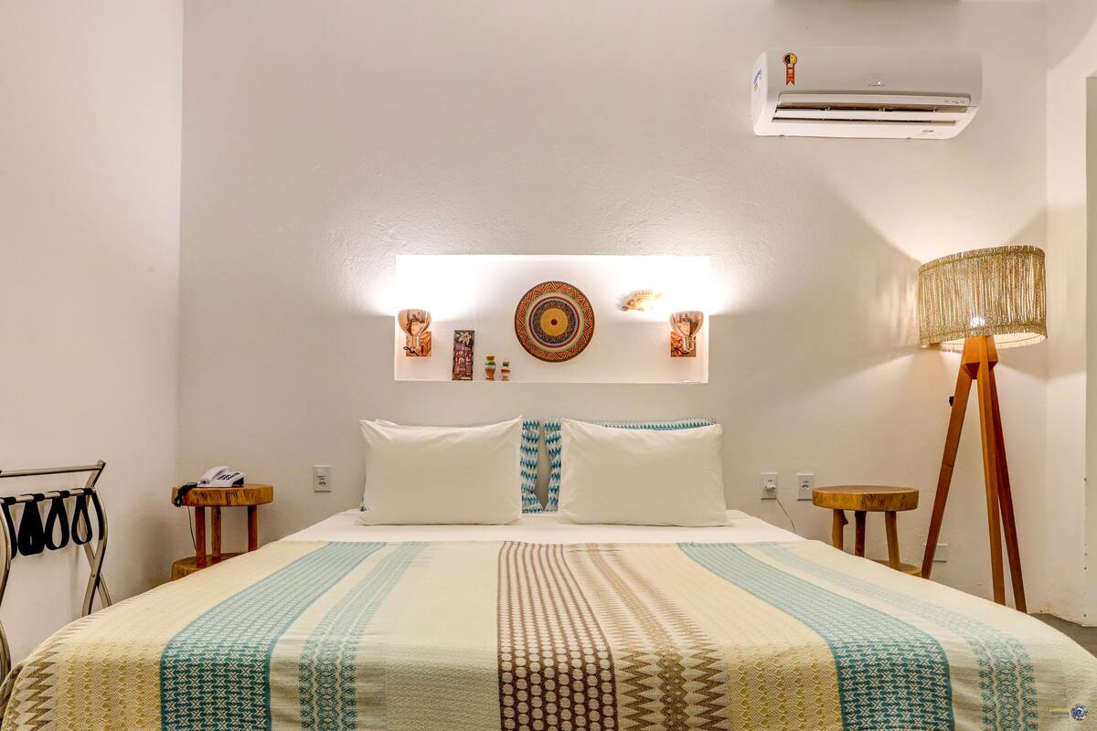Une chambre d’hôtel comprenant un lit, une table de chevet avec une lampe, un climatiseur fixé au mur et un élément décoratif mural au-dessus du lit.