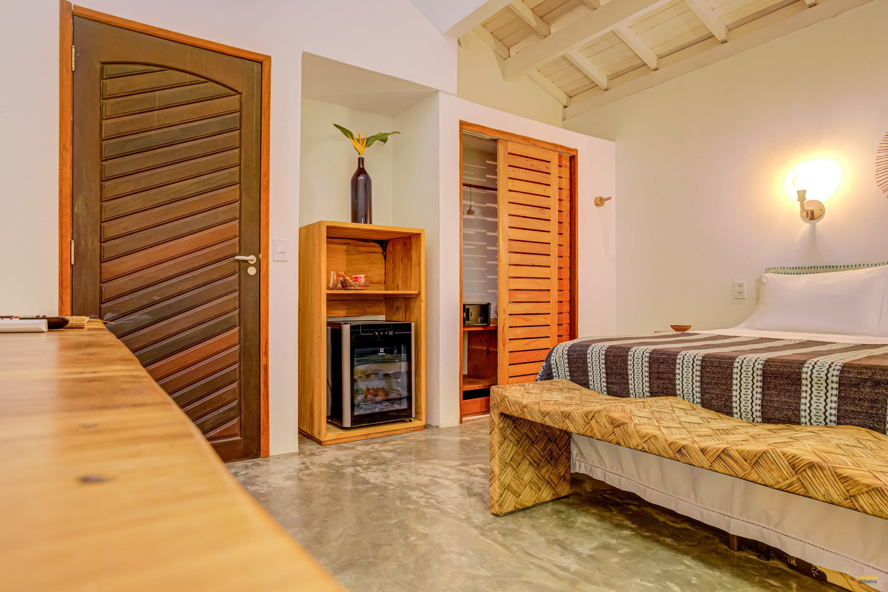 A room with a wooden door, a built-in oven, a bed with a woven bench, and a wall-mounted light fixture.