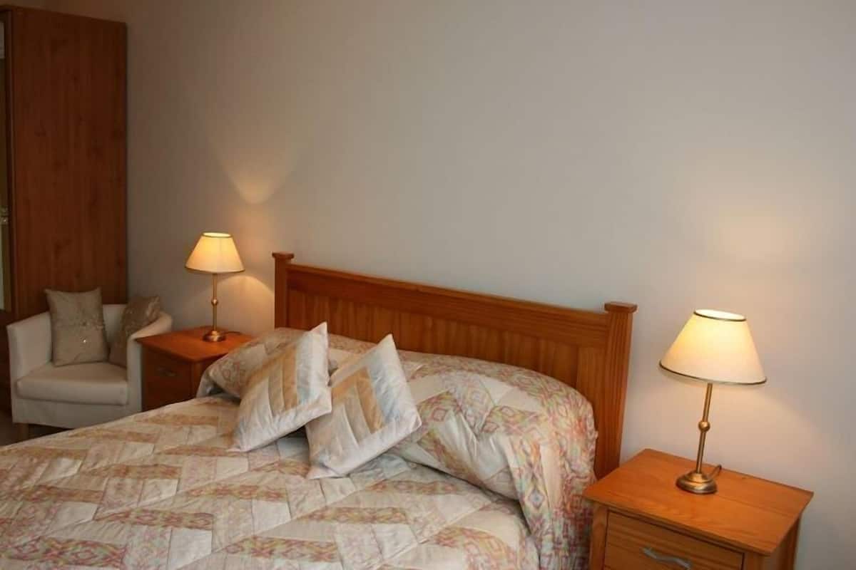 A bedroom with a wooden headboard, a bed with patterned bedding, two bedside tables with lamps, and a chair.