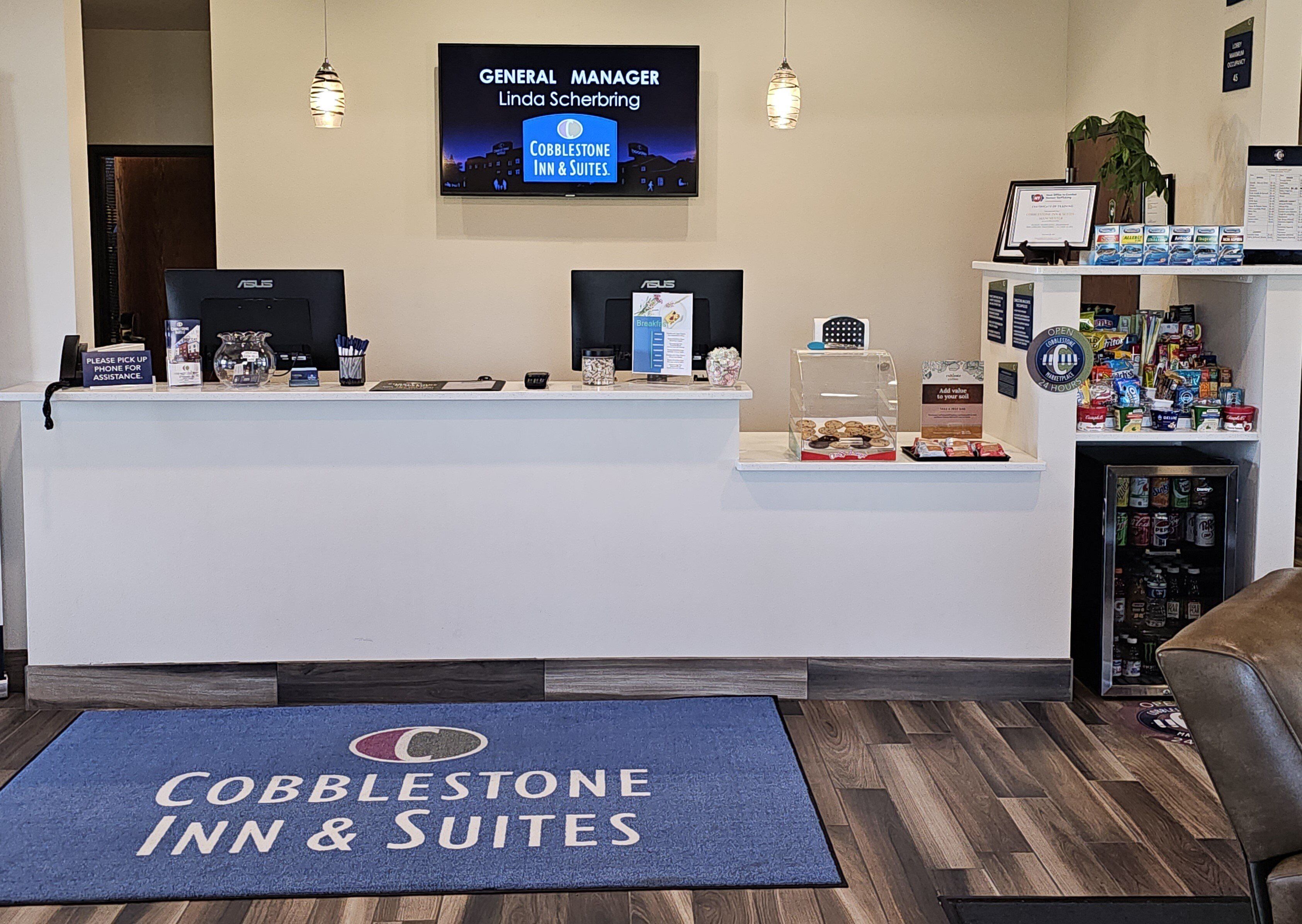 Un mostrador de recepción en Cobblestone Inn & Suites con un cartel que indica Gerente General Linda Scherbring, una computadora, una estación de café y una barra de snacks.