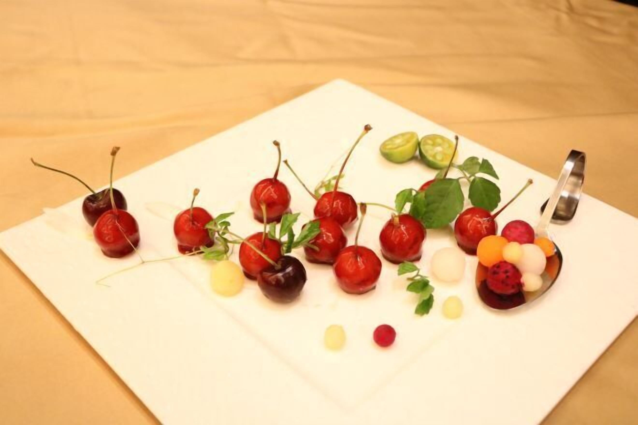 A white cutting board with cherries, lime slices, and a spoon on a wooden surface.