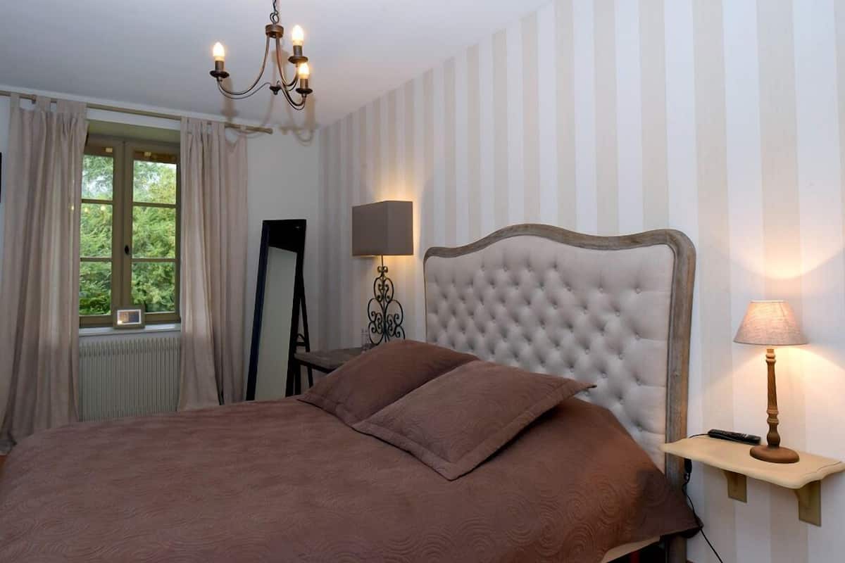 A bedroom with a tufted headboard, a bedside lamp, a mirror, and a window with curtains.