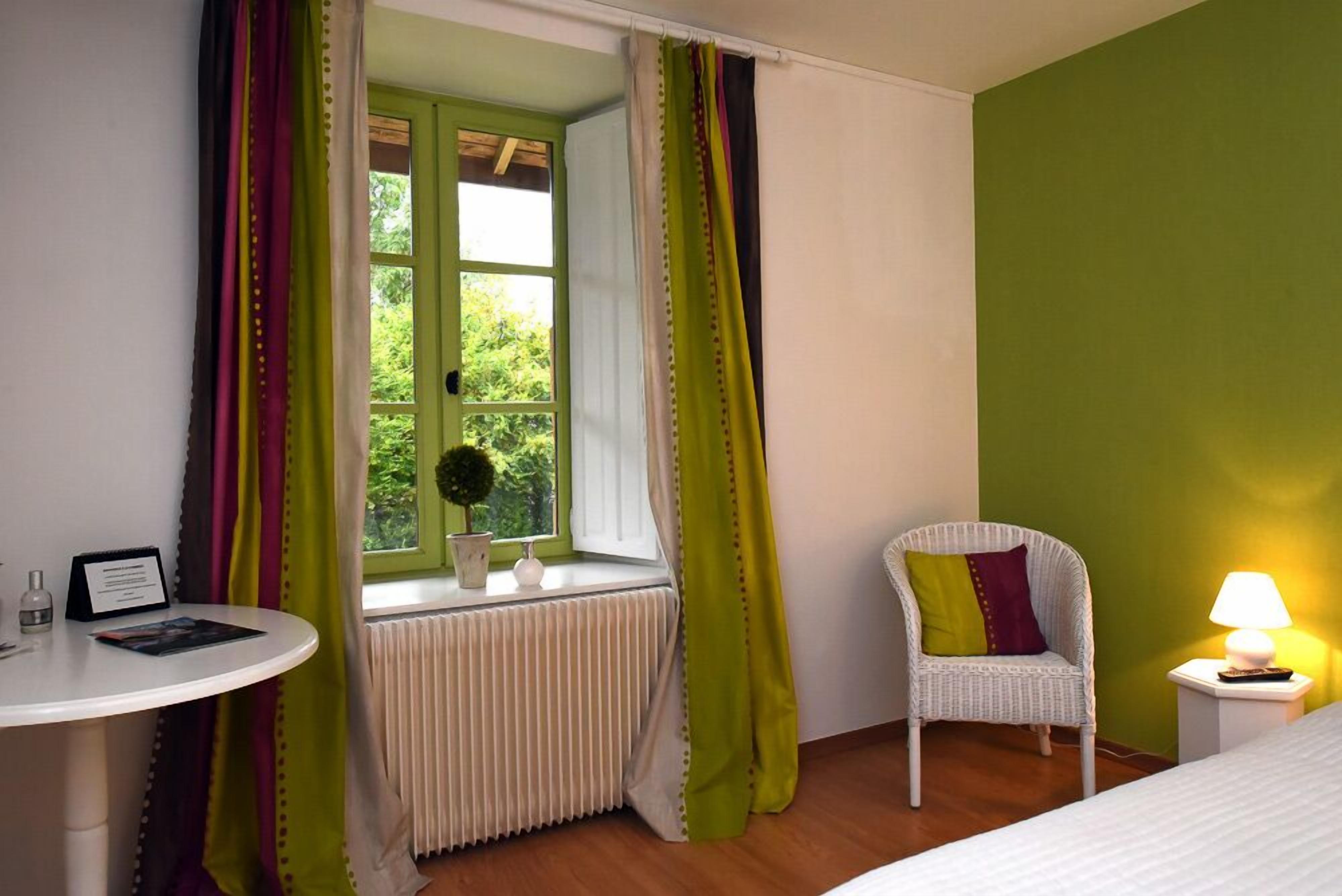 Una habitación con ventana, un radiador, una silla, una mesita de noche con lámpara y una pequeña mesa con un cartel y una botella.