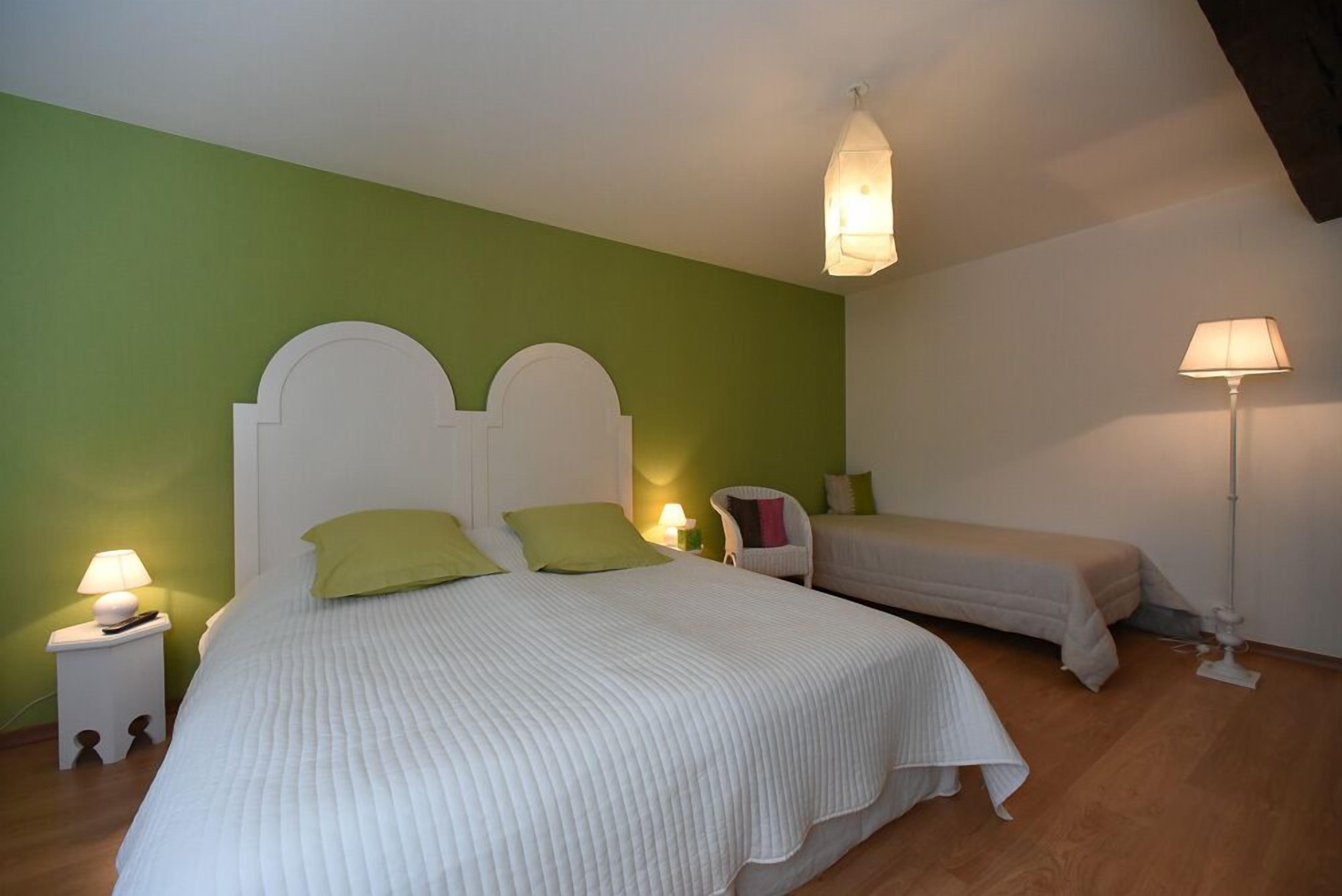 A bedroom with a large bed and a smaller bed, both with white headboards and green pillows. There are bedside lamps on nightstands, and a floor lamp in the corner.
