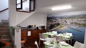 A dining area with tables set for four, a kitchen area with a view of a cityscape, and a staircase leading to an upper level. - Hostal Vendaval (Ortigueira)