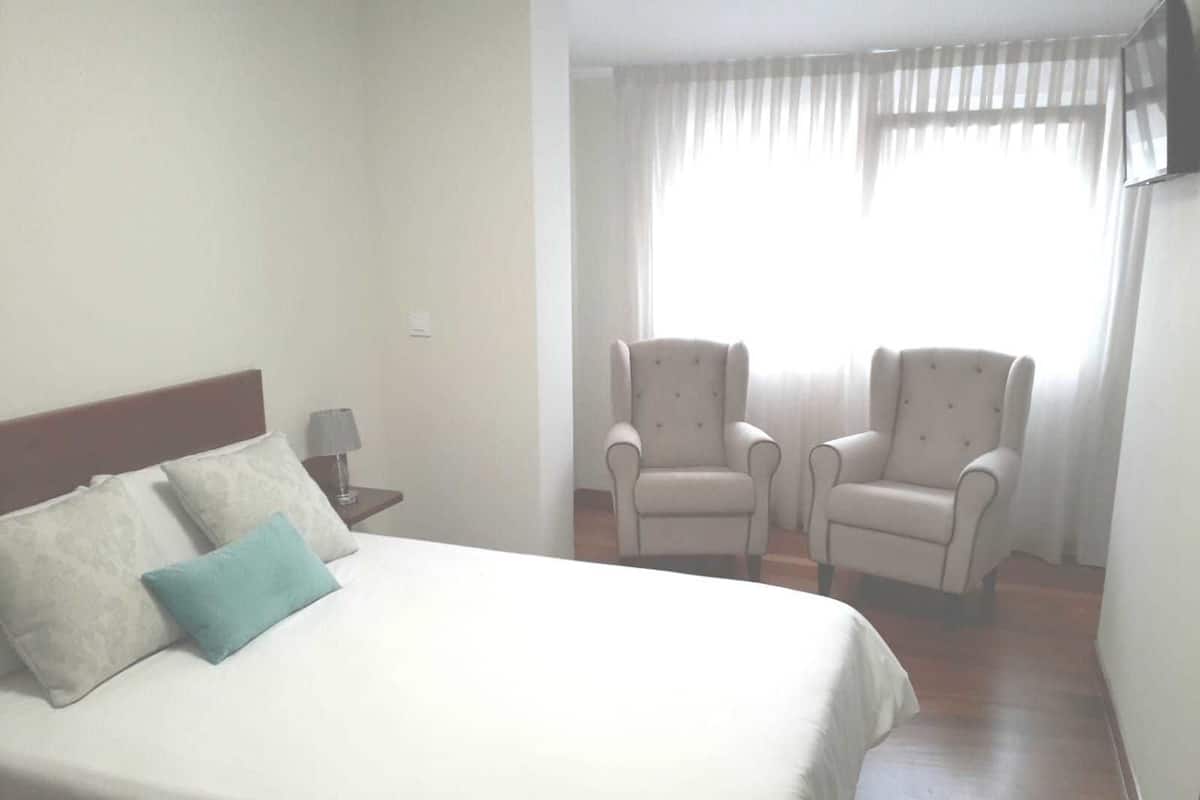 A hotel room with a bed, two armchairs, a nightstand with a lamp, and a window with curtains.