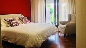 A bedroom with a large bed, two patterned pillows, a chair, and a view of greenery through a window. - Hostal Vendaval (Ortigueira)
