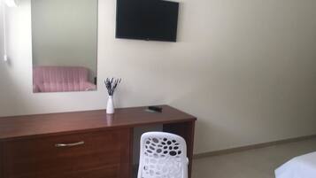 Un dormitorio con un aparador de madera, una silla de plástico blanco, una cama, un televisor de pantalla plana montado en la pared y un espejo sobre el aparador.