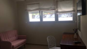 A room with a pink sofa, a white chair, a wooden desk, a TV, and a window with a view of a waterfall.