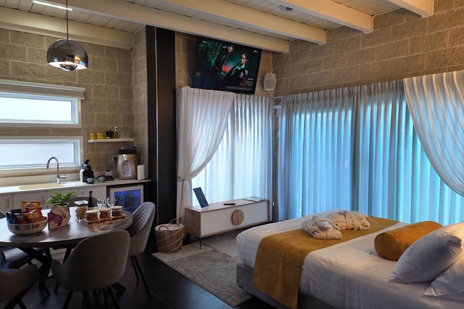 A hotel room with a bed, a desk with a computer, a window with curtains, and a television on the wall.