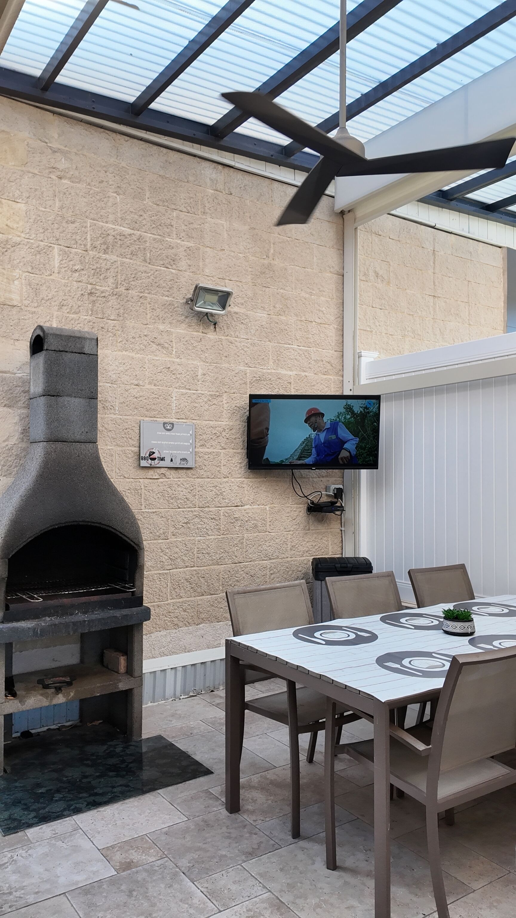An outdoor patio with a stone wall, a TV mounted on the wall, a grill, and a dining table with chairs.
