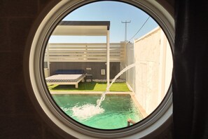A circular window view of a pool area with a bed and a small table, and a water feature. - Ahuzat Maya (Ma'ale Yosef)
