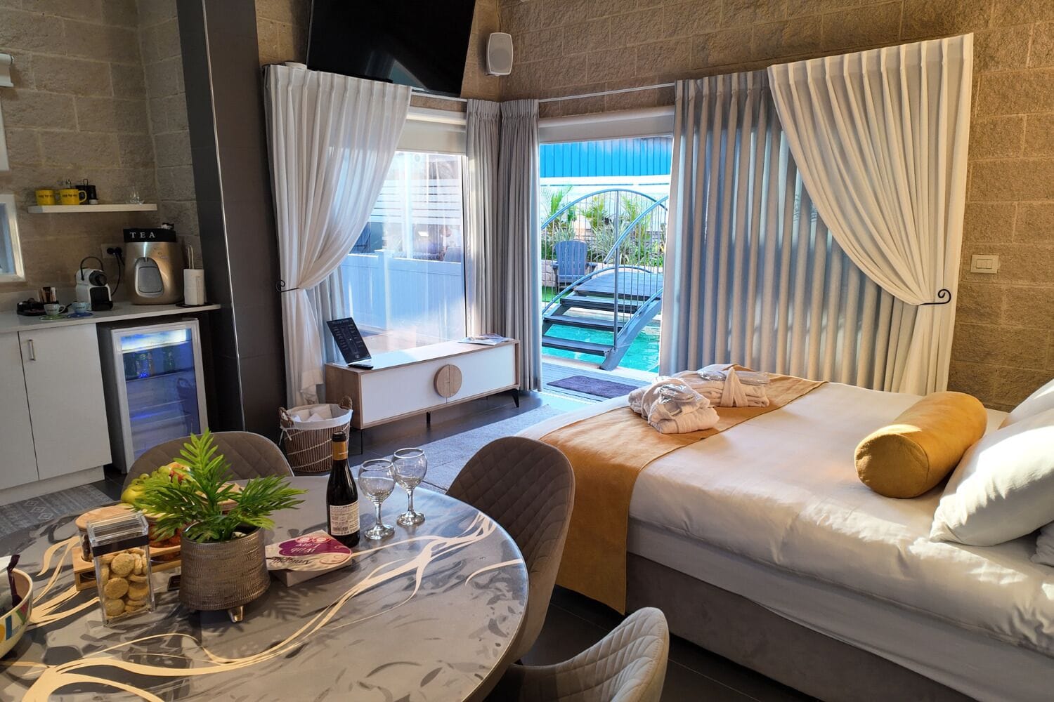 Et hotelværelse med seng, et spisebord med en plante, et minibar og udsigt til en pool.