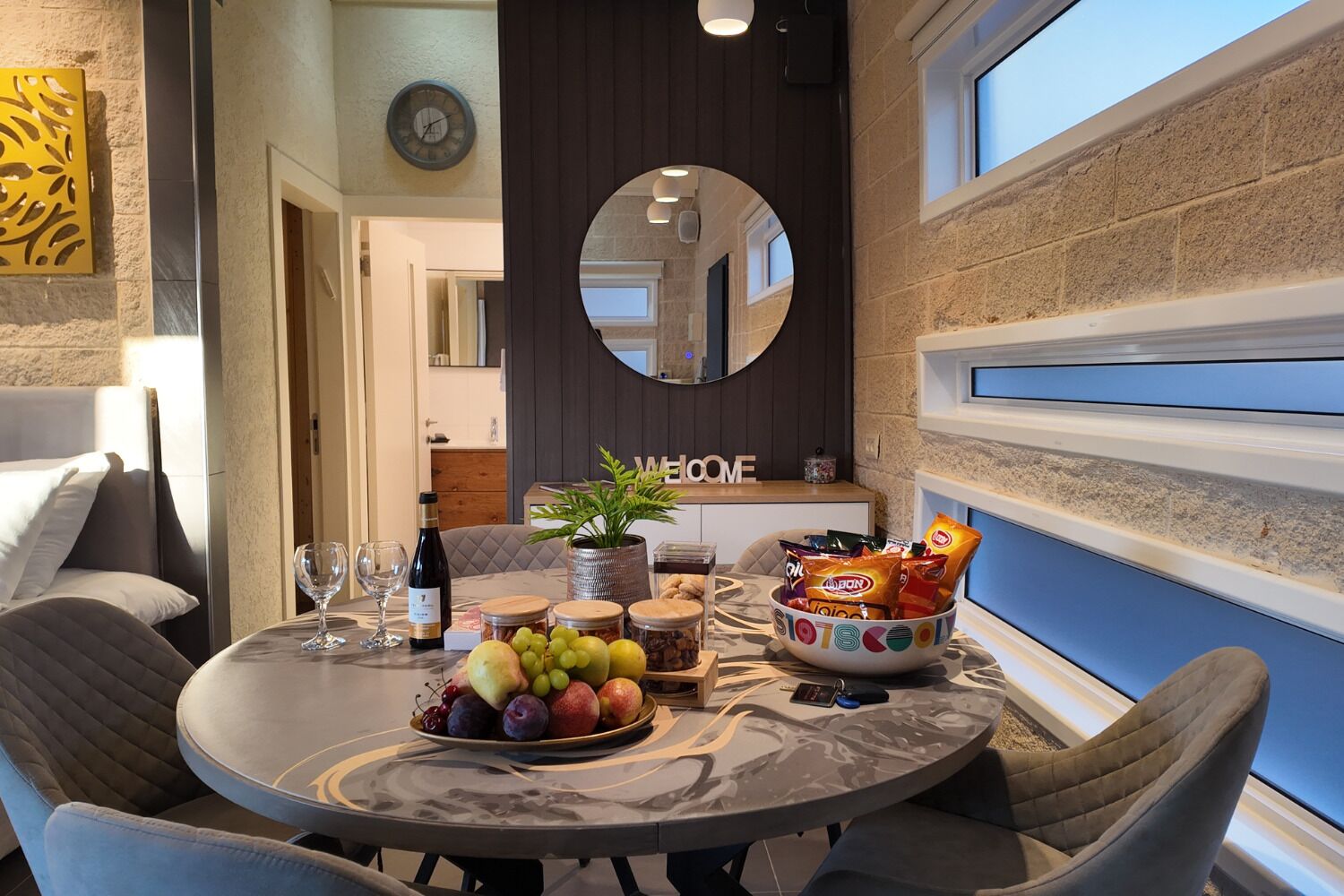 Een eethoek met een ronde tafel, stoelen, een schaal fruit, een fles wijn en een spiegel aan de muur.
