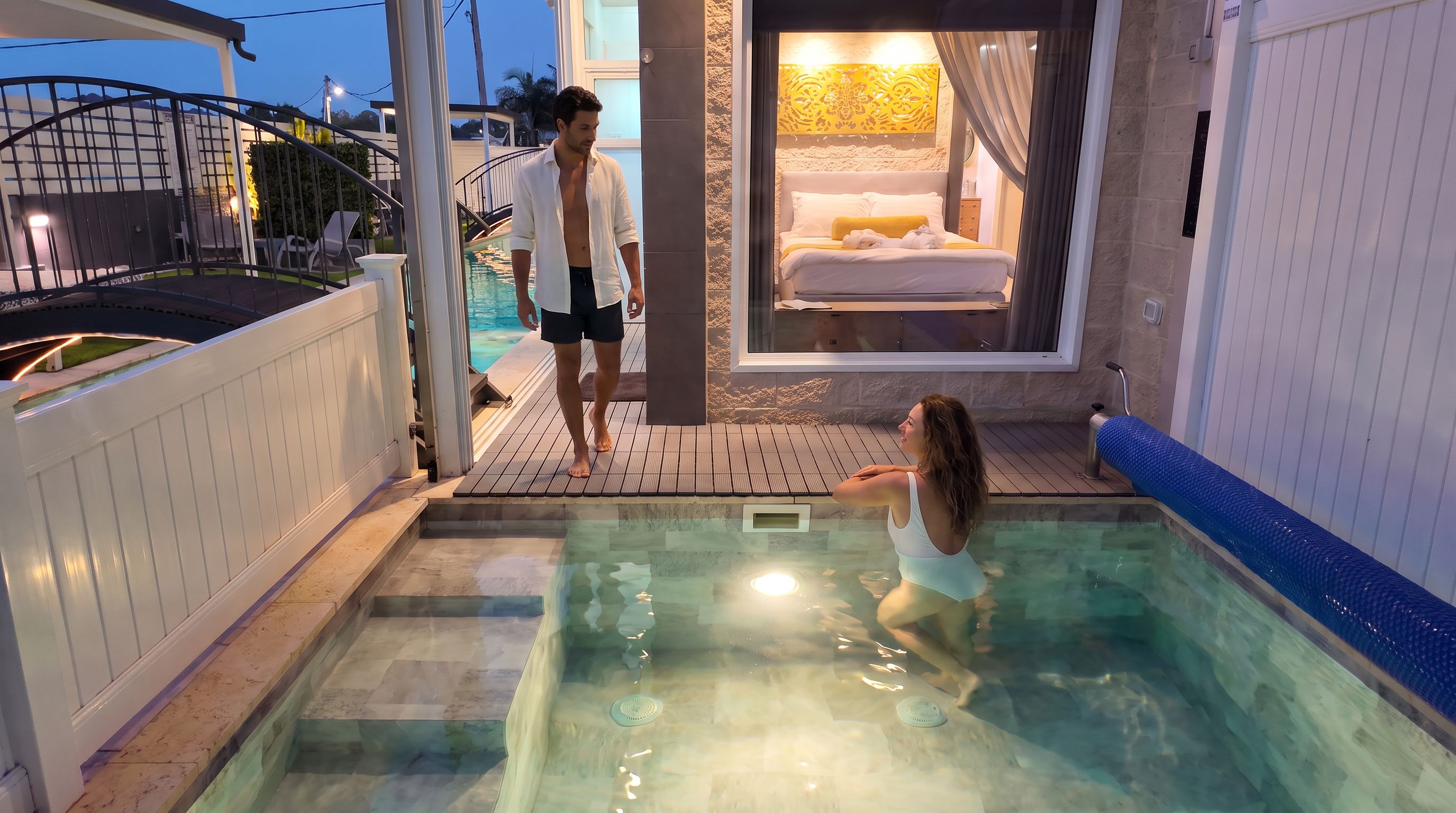 Una piscina con fondo de cristal, un dormitorio con cama y cortinas, y vista a un edificio en el fondo.