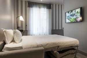 A neatly arranged hotel room with a bed, a bedside lamp, a TV mounted on the wall, and a window with curtains.