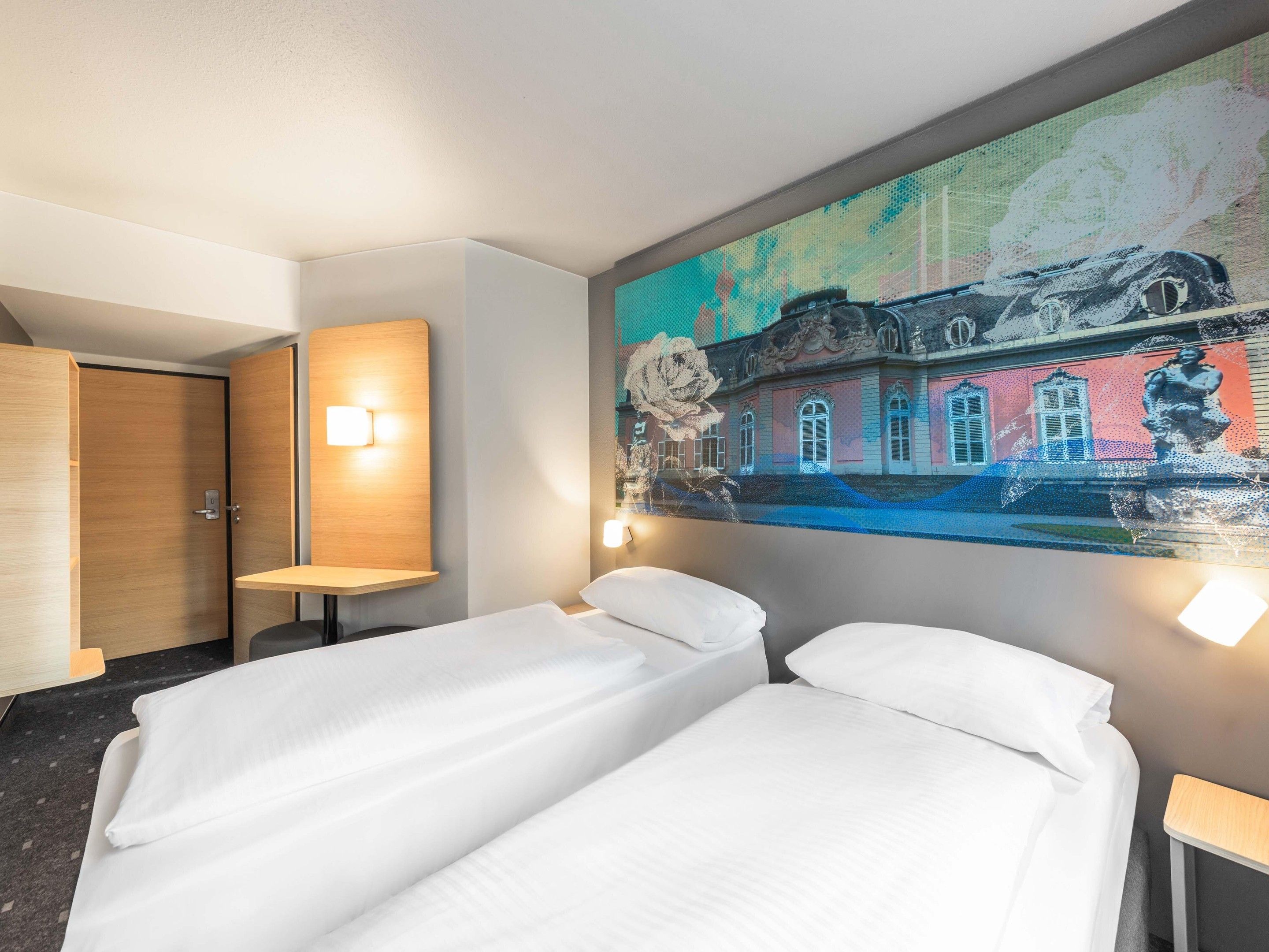 A hotel room with two single beds, a wooden wardrobe, a small table, and a wall-mounted light.